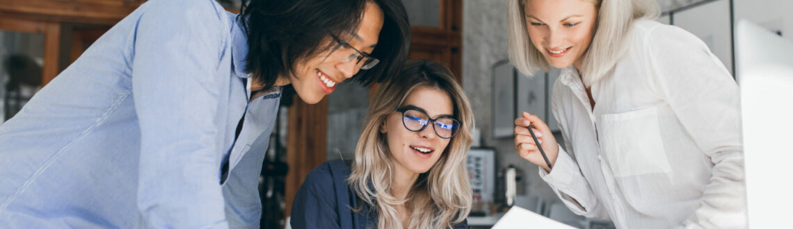 Beautiful blonde girl in glasses showing report to asian colleague which standing beside her table. Indoor portrait of cheerful international students analysing exam tasks.